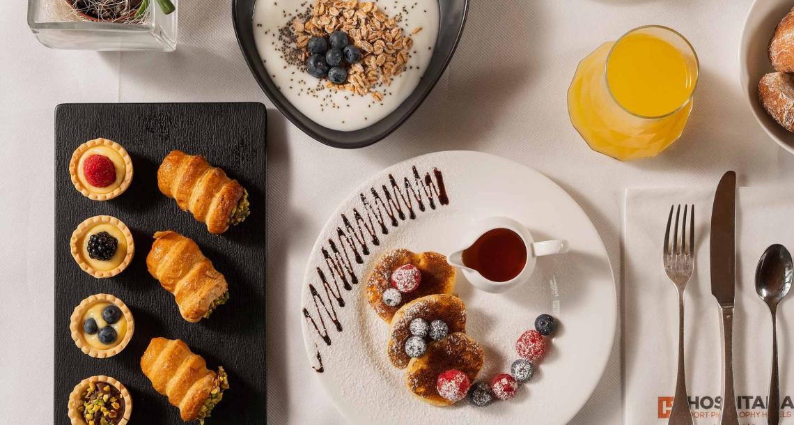 Colazione gourmet con dolci, yogurt, frutta, succo d'arancia e posate su tovagliolo.