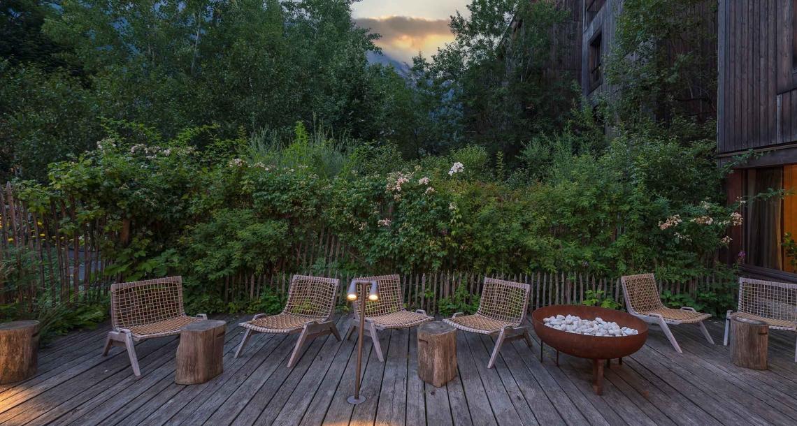 Terrazza con sedie intrecciate, tavolini in legno e braciere, circondata da vegetazione.