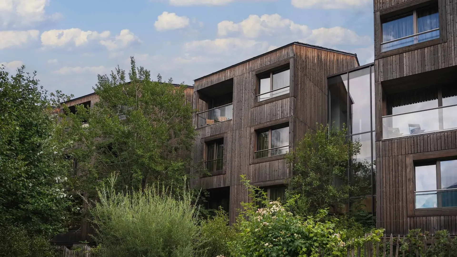 Edificio moderno in legno circondato da alberi e vegetazione, cielo nuvoloso sullo sfondo.