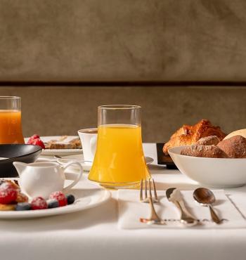 Colazione elegante con succo d'arancia, croissant e frutta fresca su tavola apparecchiata.